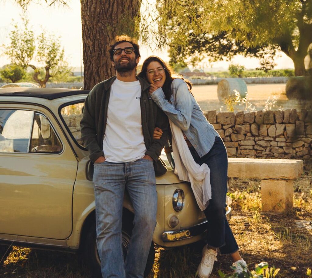 Francesco and Olga, founders of Masseria Olga, smiling and leaning on a vintage car in the Italian countryside