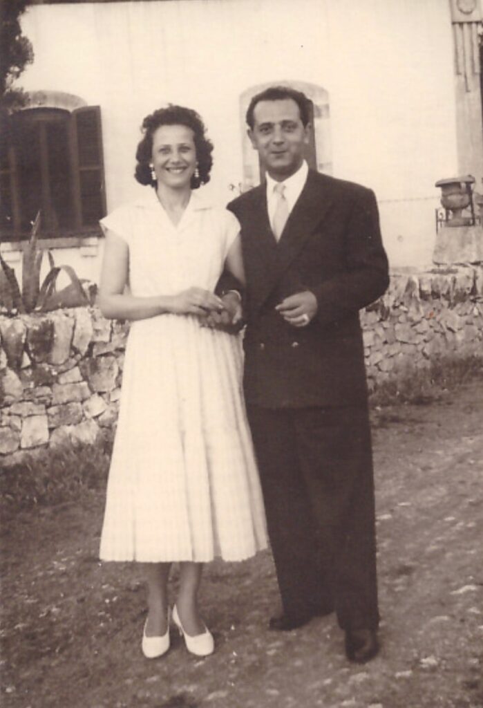 Olga wearing a light dress standing next to Vito in suit in front of the stone wall of Masseria Olga, Puglia countryside