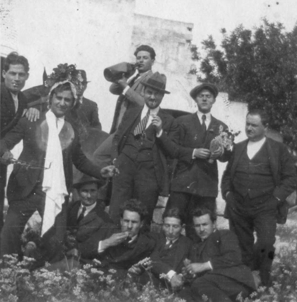 Black and white photo of men in suits, some in costume, smiling and drinking during a celebration at Masseria Olga